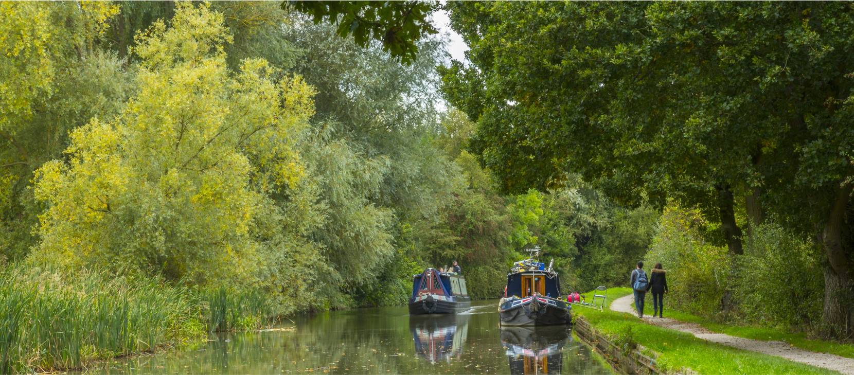 Two canal boats on the Grand Union Canal and two people walking down the towpath